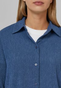 Chemise bleue texturée avec un col, boutonnage à l'avant et un subtil motif de tissu. Équipée de boutons foncés et d'une chemise intérieure blanche.
