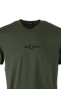 Camiseta de algodón verde oliva con cuello redondo, presenta un logotipo bordado en negro y un diseño de laurel en el centro. Mangas cortas, textura suave.