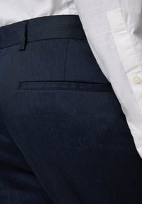 Marinblå byxor med en slät tygfinish, som har en subtil textur, en platt midja och en enstaka bakficka. Vit skjorta delvis synlig.