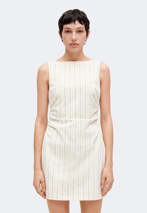 Jeune femme aux cheveux courts et foncés portant une robe blanche ajustée sans manches avec de fines rayures verticales, debout devant un fond uni.