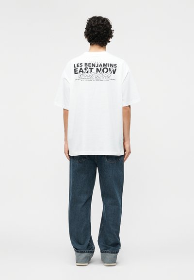 Άτομο με σγουρά σκούρα μαλλιά που φοράει λευκό, φαρδύ T-shirt με μαύρο κείμενο και μπλε φαρδιά τζιν, στέκεται με την πλάτη γυρισμένη σε απλό φόντο.