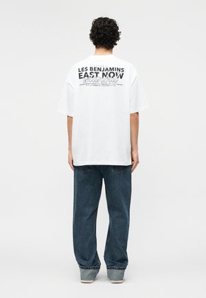 Person with curly dark hair wearing a white oversized T-shirt with black text and blue loose jeans, standing facing away on a plain background.