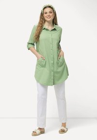 Camicia verde chiaro con bottoni e tasche, maniche arrotolate, abbinata a pantaloni bianchi e sandali beige. Capelli acconciati con un fermaglio coordinato.
