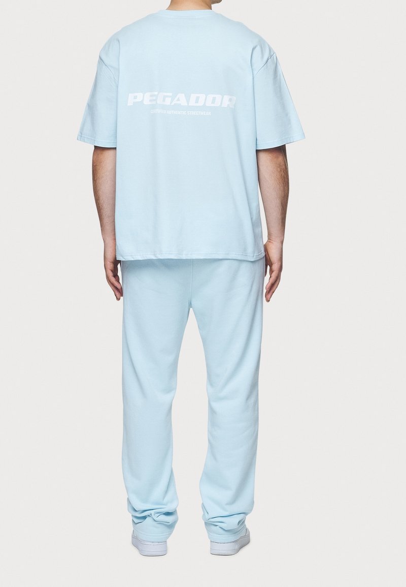 T-shirt en coton oversize bleu clair avec du texte blanc "PEGADOR" au dos, associé à un pantalon de survêtement bleu clair assorti et des baskets blanches.