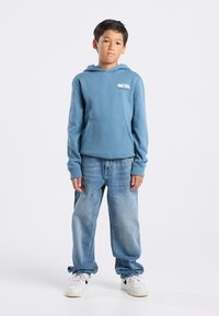 Sweatshirt à capuche bleue avec poche avant, texture douce. Associé à un jean bleu clair ample et des baskets blanches avec des accents noirs.