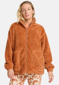 Veste zippée orange duveteuse avec deux poches avant et une texture épaisse et moelleuse. Dispose d'une fermeture éclair contrastante et d'un col arrondi.