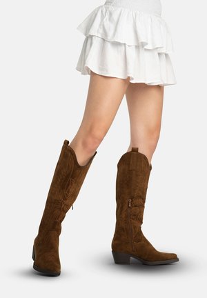 Kvinde iført brune knæhøje cowboystøvler i ruskind med broderi og en hvid lagdelt mininederdel, stående på en hvid baggrund.