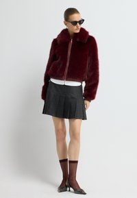 Burgundská umělá kožešinová bomber bunda s šálovým límcem, kombinovaná s plisovanou černou sukní, průhlednými burgundskými ponožkami a špičatými lodičkami.