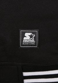 Zwarte stof met een wit rechthoekig label waarop "STARTER BLACK LABEL" staat en een gestileerd sterlogo. Bevat horizontale zwart-witte strepen.