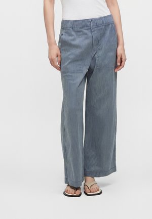 Persoană purtând pantaloni largi cu dungi verticale albastre și albe, talie elastică și sandale, stând în fața unui fundal simplu.