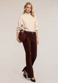Suéter beige acanalado con cuello alto, combinado con pantalones de pana marrón oscuro. Sosteniendo un bolso burdeos, llevando tacones altos negros.
