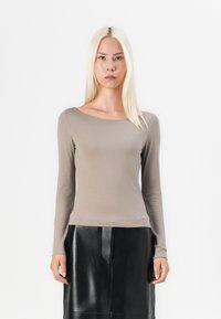 Top lung cu mânecă lungă de culoare taupe, cu decolteu tip barcă, realizat dintr-un material moale, cu un accent subtil al logo-ului în partea de jos. Asortat cu o fustă neagră din piele.