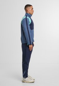 Granatowy i turkusowy dres wykonany z lekkiego materiału; posiada zamki, wysoki kołnierz i elastyczne mankiety; noszony z kremowymi trampkami.