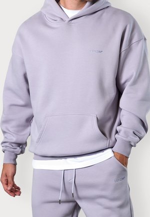 Hombre con sudadera morado claro y pantalones de chándal a juego con una camiseta blanca debajo, manos relajadas, logo visible de ESTIIM en el pecho y los pantalones.