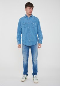 Camicia di jeans azzurro chiaro con due tasche sul petto, yoke appuntito e colletto button-down; abbinata a jeans azzurro chiaro e sneaker bianche.