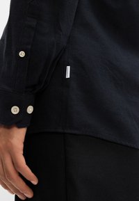 Cămașă cu mânecă lungă de culoare bleumarin, realizată dintr-un material moale, cu manșete cu nasturi și un mic etichetă albă de brand aproape de tiv, asociată cu pantaloni închiși la culoare.