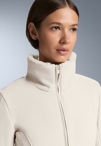 Jasno-beżowa kurtka zapinana na zamek z wysokim kołnierzem i miękką, białą wyściółką z polaru. Gładka faktura i minimalistyczny design bez widocznych elementów metalowych.