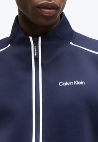 Κλείσιμο εικόνας ατόμου που φοράει ναυτικό μπλε μπουφάν Calvin Klein με λευκό φερμουάρ και λογότυπο στο στήθος.