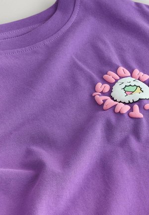 Tricou mov din bumbac, cu un personaj de desen animat alb imprimat, având o gură în formă de curcubeu, înconjurat de text roz. Textură netedă.