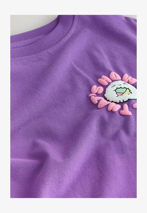 Fialové bavlnené tričko s potlačou bielej kreslenej postavičky s dúhovými ústami, obklopenej ružovým textom. Hladká textúra.