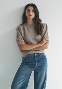 Kobieta z długimi ciemnymi włosami, ubrana w beżowy sweter z krótkim rękawem i niebieskie jeansy, stojąca z ramionami skrzyżowanymi na tle białej ściany.