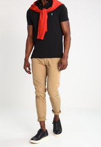 Svart polo shirt med blå logotyp, röd tröja knuten runt nacken, beige chinos med uppviken byxben och svarta sneakers med vita sulor.