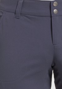 Närbild av midjebandet på marinblå byxor med bälteshällor, framfickor och två silverfärgade tryckknappar på högra sidan.