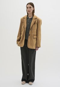 Tan suède blazer over een grijs gebreid polo, gecombineerd met losse zwarte pinstripe broeken en lichte schoenen, wat een gelaagd uiterlijk creëert.
