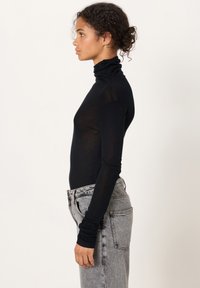 Mladá žena s kudrnatými vlasy, oblečená v černém roláku a šedých džínách s vysokým pasem, stojící v profilu na jednobarevném pozadí.