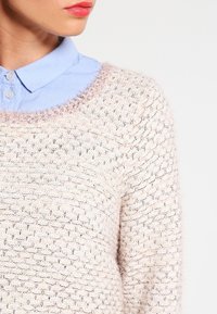 Joven vistiendo una camisa color azul pastel con cuello debajo de un suéter de punto ligero color rosa claro, frente a un fondo blanco puro.