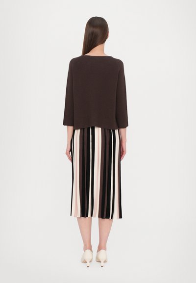 Pull court brun à manches 3/4 porté sur une jupe plissée avec des rayures verticales noires, roses et crème. Chaussures à talons blanches.