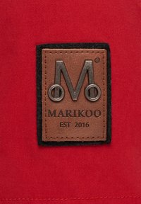 Smeđa kožna oznaka s srebrnim logotipom "M" i tekstom "MARIKOO EST 2016", postavljena na crvenu tkaninu s crnim rubom.