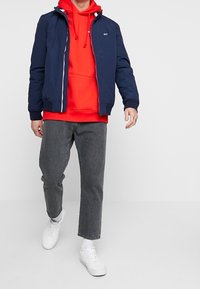 Marinblå bomberjacka med en röd huvtröja under, grå tapered byxor och vita sneakers. Fokusera på lager och kontrast.