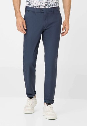 Pantaloni blu navy con una vestibilità su misura, dotati di chiusura a bottone, tasche laterali e una texture liscia, abbinati a sneakers bianche.