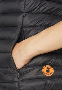 Chaleco puffer negro con un bolsillo con cremallera visible y un logo circular naranja con la silueta de un pato, sobre un fondo de textura acolchada.