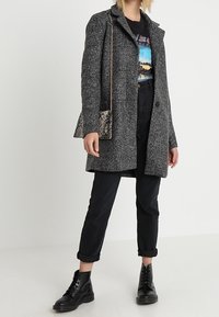 Grijze jas van een wolmix met een klassieke kraag, gecombineerd met een grafisch t-shirt, zwarte omgeslagen broekspijpen en zwarte veterlaarzen. Handtas met slangenprint.