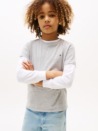 T-shirt grigio a maniche corte sovrapposto a una camicia bianca a maniche lunghe, con un dettaglio del logo. Jeans in denim blu chiaro. Capelli ricci. Sfondo neutro.