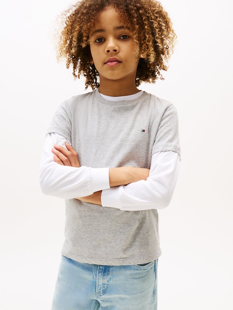 T-shirt grigio a maniche corte sovrapposto a una camicia bianca a maniche lunghe, con un dettaglio del logo. Jeans in denim blu chiaro. Capelli ricci. Sfondo neutro.