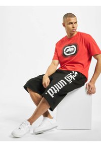 Rød bomulls T-skjorte med et sirkulært grafisk logo, kombinert med svarte knelange shorts med hvit tekst, og hvite sneakers på en hvit pedestal.