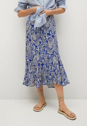 Femme portant une jupe midi bleue à motif cachemire, une chemise bleu clair rayée et des sandales plates beiges avec des brides à la cheville, debout devant un mur blanc uni.