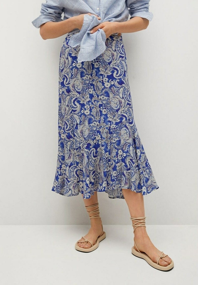 Femme portant une jupe midi bleue à motif cachemire, une chemise bleu clair rayée et des sandales plates beiges avec des brides à la cheville, debout devant un mur blanc uni.