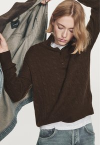 Donna con capelli biondi corti che indossa un maglione a trecce marrone scuro sopra una camicia grigia chiara, sollevando una giacca grigia con etichetta "Massimo Dutti".