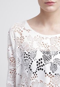Weißes Strickoberteil mit einem spitzenähnlichen Muster, das unterschiedliche Lochgrößen und florale Formen aufweist und Transparenz sowie strukturierte Details präsentiert.