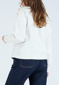 Weißes Sweatshirt mit lässiger Passform, langen Ärmeln und goldenen Knopfakzenten an der Schulter. Kombiniert mit dunkelblauen Jeans.