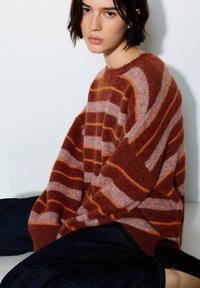 Oversized trui met roodbruine en roze strepen, gemaakt van zacht, pluizig materiaal. Heeft brede mouwen en een ronde hals, gecombineerd met een donkere spijkerbroek.
