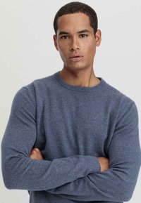 Blauer Wollpullover mit Rundhalsausschnitt, gerippten Bündchen und weicher Textur, getragen von einem Model mit verschränkten Armen, neutralem Hintergrund.