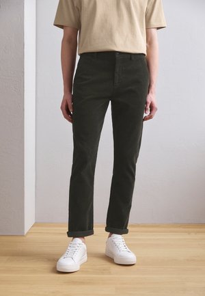 Persona con maglietta beige, pantaloni scuri arrotolati e sneakers bianche, in piedi su un pavimento in legno chiaro davanti a una parete semplice.