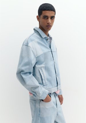 Junger Mann trägt eine hellblaue Patchwork-Jeansjacke und Jeans, steht mit einer Hand in der Tasche vor einem weißen einfarbigen Hintergrund.