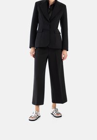 Blazer nero su misura con rever a picco, abbinato a pantaloni neri a gamba larga e sandali metallici argento con molteplici cinturini e suole nere.