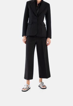 Blazer nero su misura con rever a picco, abbinato a pantaloni neri a gamba larga e sandali metallici argento con molteplici cinturini e suole nere.